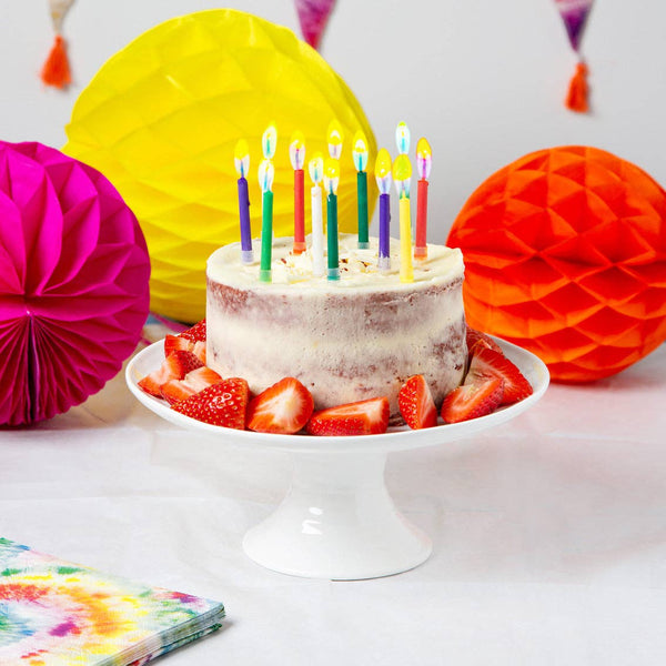 Birthday cake with lit candles on a white plate, surrounded by colorful paper decorations.