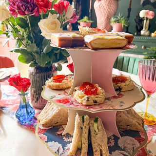 Three-tiered cake stand with finger sandwiches, scones, and pastries on a floral tablecloth.