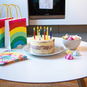 Birthday cake with lit candles on a table next to a rainbow-colored gift bag.