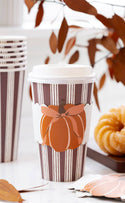 A white and brown striped paper coffee cup with a die-cut autumn pumpkin graphic, shown on a tabletop with stacked cups, fall leaves, and a small pumpkin prop.
