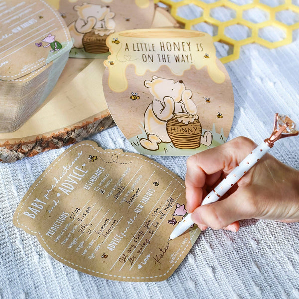 Baby shower invitation with cartoon bear and honey pot design, held by a hand with a pen.