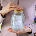 Person holding a glass jar with a gold lid, possibly containing honey, against a neutral background.