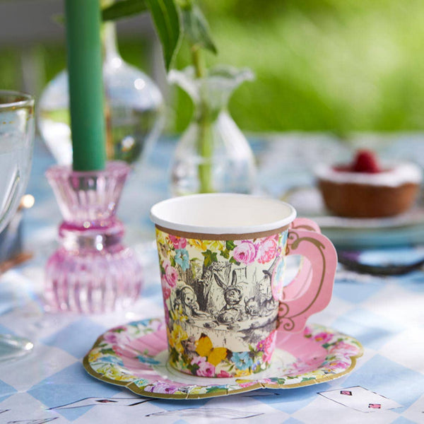 Decorative teacup and saucer on a table with a floral pattern, surrounded by other tableware and plants.