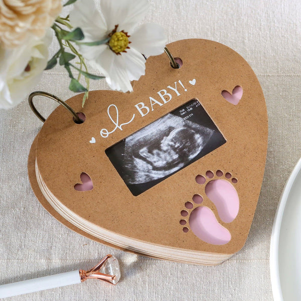 Heart-shaped photo album with ultrasound inside, surrounded by flowers on a light surface