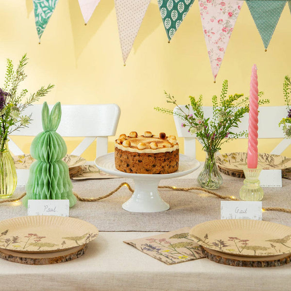 Table setting with a cake, bunny decorations, and floral arrangements against a yellow wall with bunting.