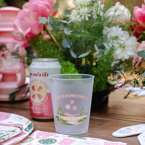 Frosted cup with 'Bachelorette' design, Pina Colada Drndrift can, and floral arrangement on a table.