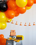 Decorative setup with orange, yellow, and black balloons, traffic cones, and a cake on a stool.