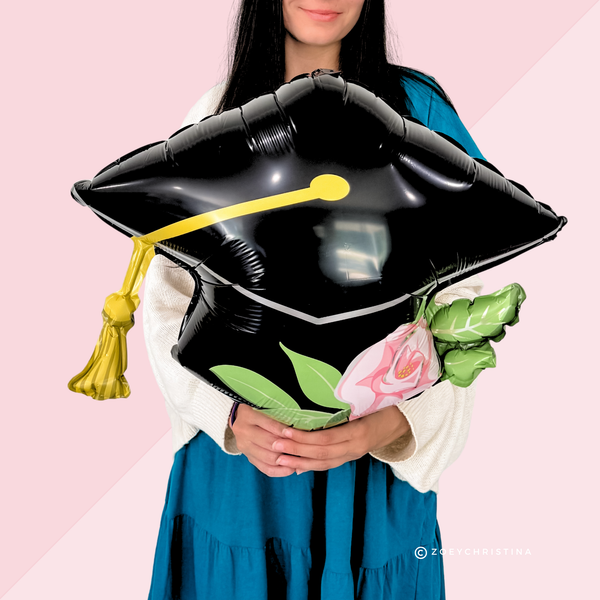 Person holding a black graduation cap balloon with a pink rose and green leaves against a pink background