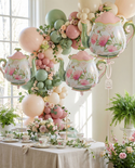 Decorative arch with floral balloons and teapot-shaped balloons in a room with plants and a table.