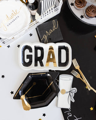 Graduation-themed party supplies including a 'GRAD' plate, black graduation cap, and gold utensils on a black surface.