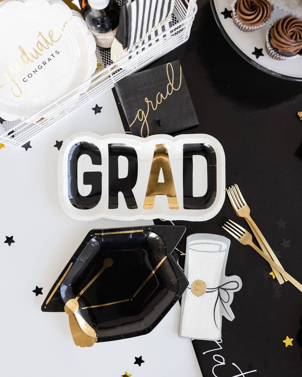 Graduation-themed party supplies including a 'GRAD' plate, black graduation cap, and gold utensils on a black surface.