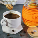 Honey being drizzled into a cup of coffee with a jar of honey and coffee beans on a dark surface.