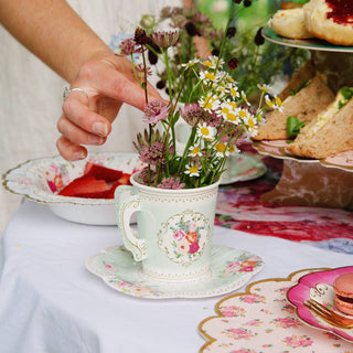 Teacup with flowers on a table set for tea, with a hand reaching in.