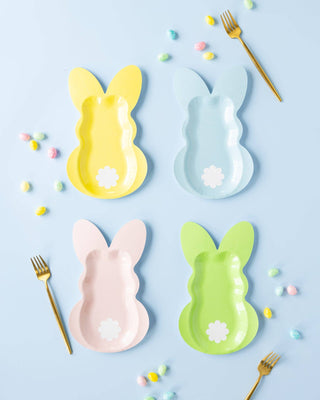 Four colorful bunny-shaped plates on a light blue background with gold forks and pastel candies.