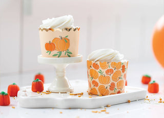 Cupcakes with pumpkin-themed wrappers on a white surface with pumpkins in the background.