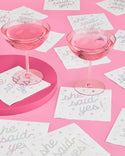Two pink cocktail glasses on a pink surface with 'she said yes' cards scattered around.