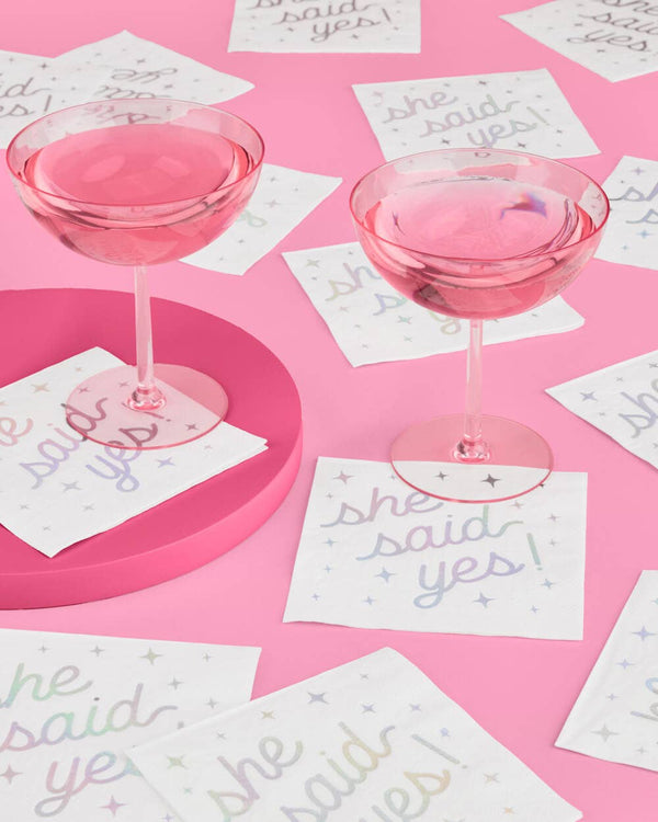 Two pink cocktail glasses on a pink surface with 'she said yes' cards scattered around.