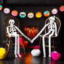 Two paper skeletons holding hands with Halloween decorations in the background.