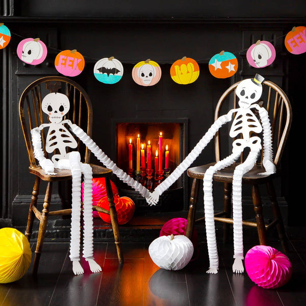 Two paper skeletons holding hands with Halloween decorations in the background.
