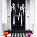 Decorative skeletons hanging on a black door with Halloween pumpkins on a checkered floor.