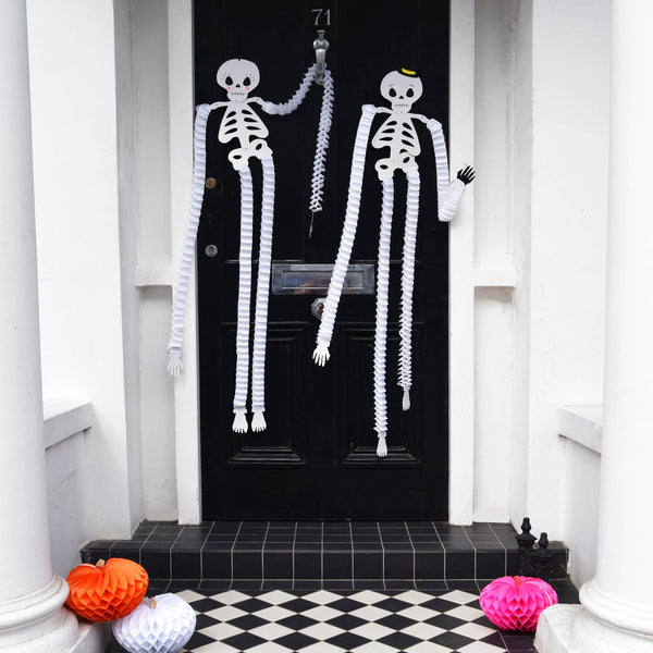 Decorative skeletons hanging on a black door with Halloween pumpkins on a checkered floor.