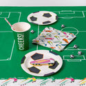 Party supplies including paper plates, cups, and napkins on a green tablecloth with soccer-themed designs.
