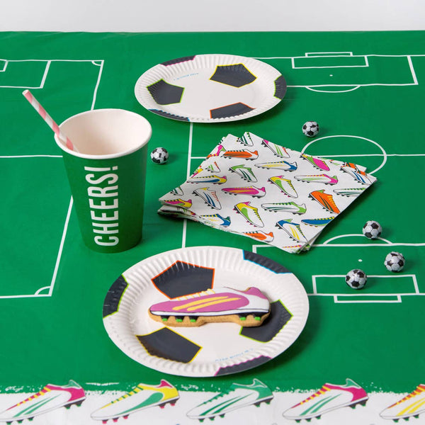 Party supplies including paper plates, cups, and napkins on a green tablecloth with soccer-themed designs.