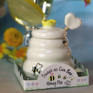 White honey pot with a yellow lid and heart-shaped dipper, featuring bumblebee designs, on a decorative stand with flowers in the background.