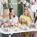 Four women enjoying a tea party with cupcakes and flowers in a garden setting.