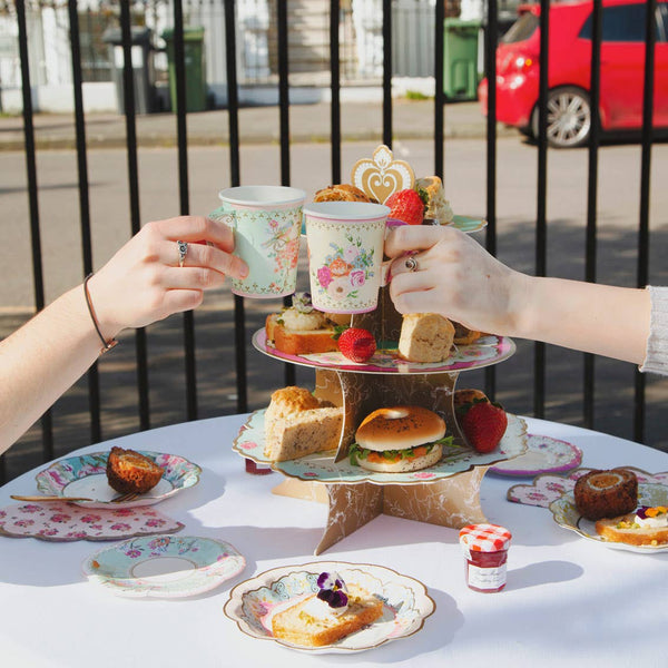 Two people holding floral teacups over a three-tiered cake stand with pastries and sandwiches outdoors.