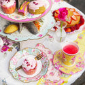 Table setting with floral plates, pink cupcake, and a cup of tea on a decorative tablecloth.