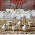 Decorative teapot and cup place card holders with floral cards on a textured surface.