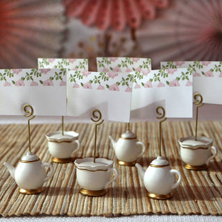 Decorative teapot and cup place card holders with floral cards on a textured surface.