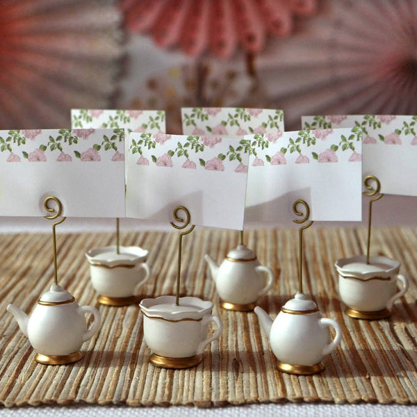 Decorative teapot and cup place card holders with floral cards on a textured surface.