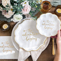 White paper plates with 'the adventure begins' text on a wooden table with flowers and candles.