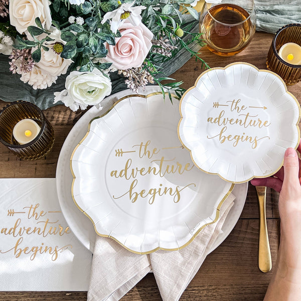 White paper plates with 'the adventure begins' text on a wooden table with flowers and candles.