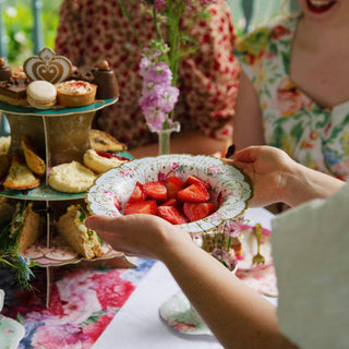 Person holding a plate of strawberries at a tea party with a tiered cake stand and floral decorations.
