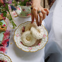 Hand with a ring holding a dessert on a decorative plate with floral patterns, surrounded by a tea set and flowers.